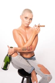 Porträt einer Frau mit rasiertem Kopf, Tattoos und zerrissenen Jeans, die eine Flasche und eine Zigarre hält.