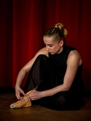 Eine junge Balletttänzerin mit blondem Pferdeschwanz bindet ihre Spitzenschuhe vor einem roten Vorhang.