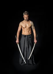 Ein muskulöser Mann mit Tattoos und grauen Haaren, der zwei Schwerter hält und einen schwarzen Hakama trägt.
