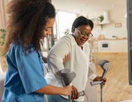 A home care worker providing steady support as an elderly woman confidently walks with her crutches