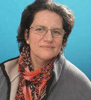 A woman with short dark hair and glasses looks toward the camera. She wears a neutral-toned jacket.