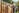 A man in overalls and a cap paints a wooden fence with a brush in a sunny backyard, surrounded by gr