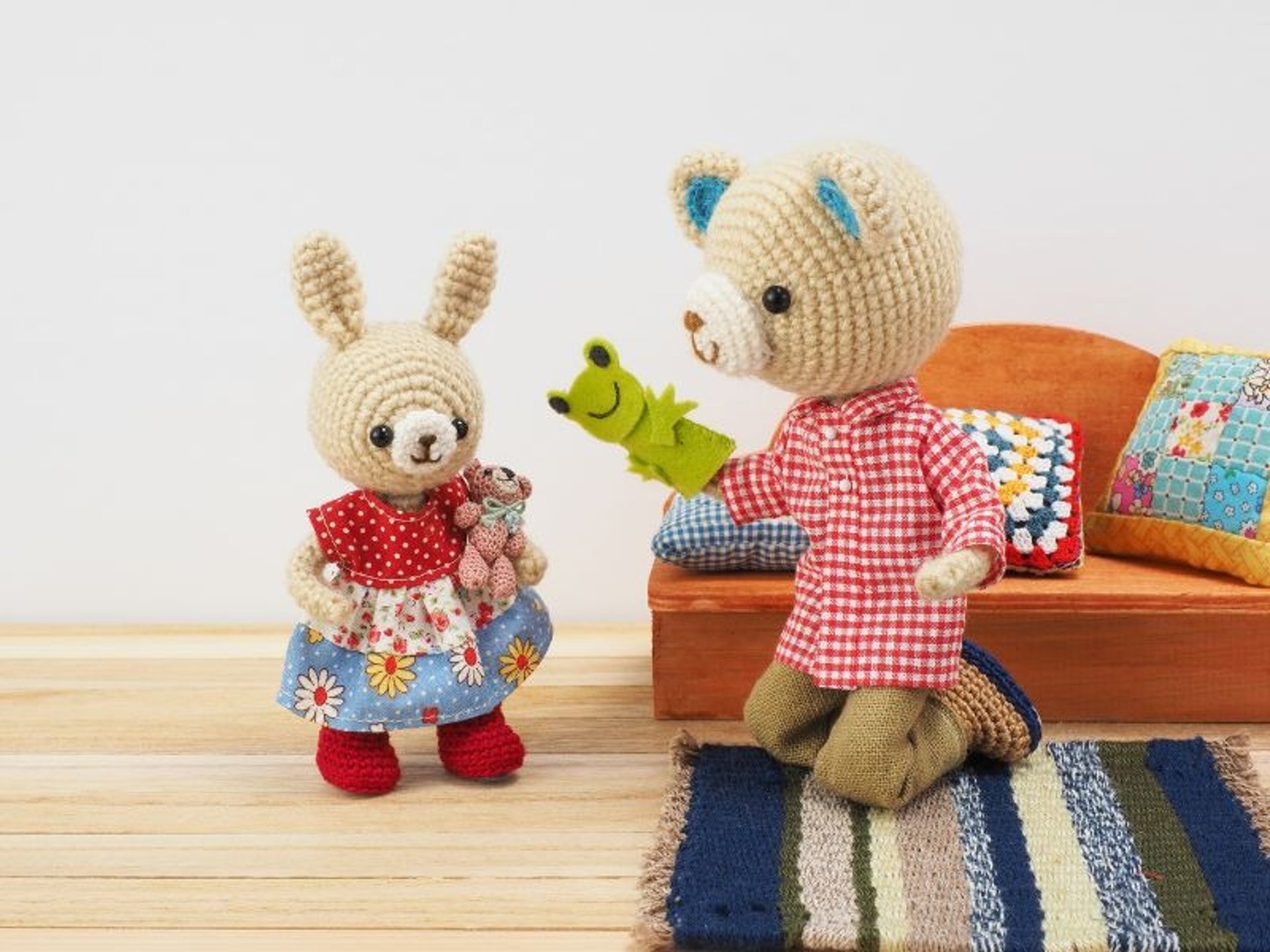 Dos muñecos amigurumi de oso y conejo con vestimenta de colores en una escena con sillón naranja con cojines