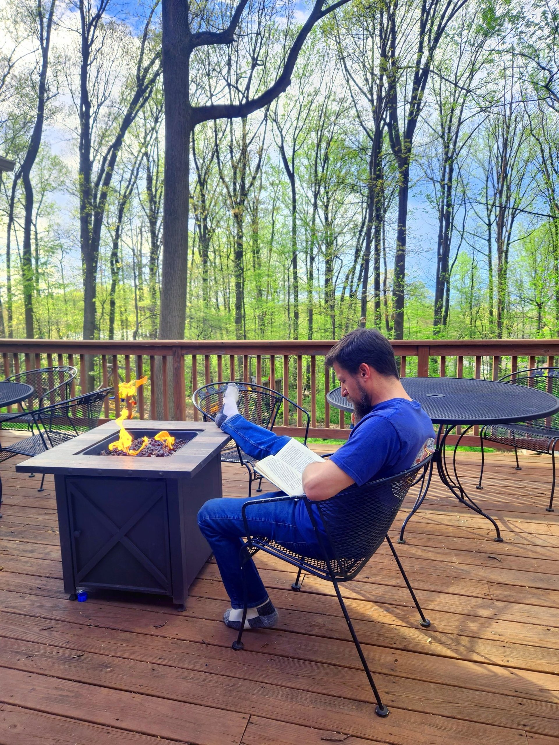 Donald Owens Sits Reading on the deck by the firepit in the woods