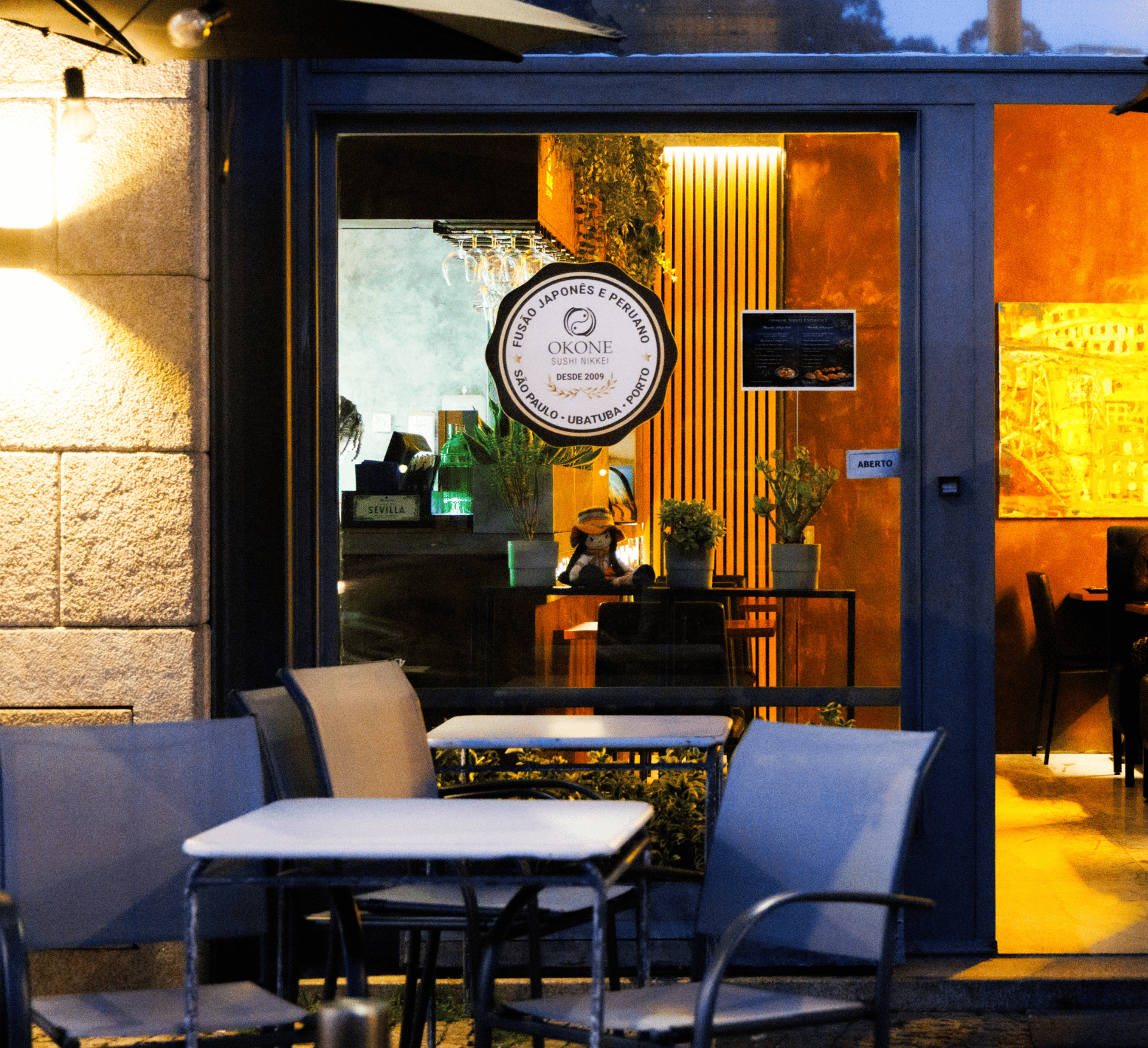 Entrada do Okone Sushi no Porto com mesas externas, fachada de vidro e logótipo iluminado durante o entardecer.