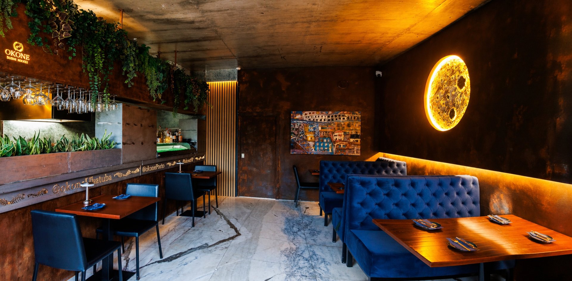 Interior do Okone Sushi Nikkei no Porto com sofás azuis, balcão de madeira, iluminação suave e decoração com plantas