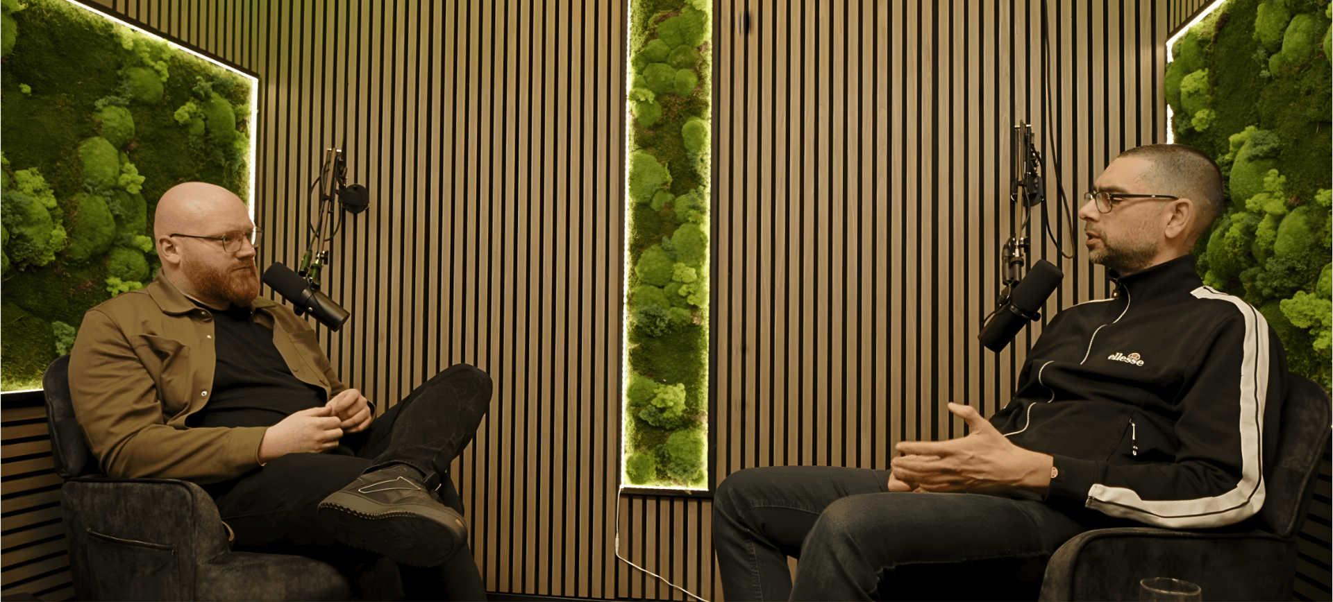 Zwei Personen sitzen sich gegenüber vor einem Mikrofon im Hintergrund sind Moosbilder zu erkennen
