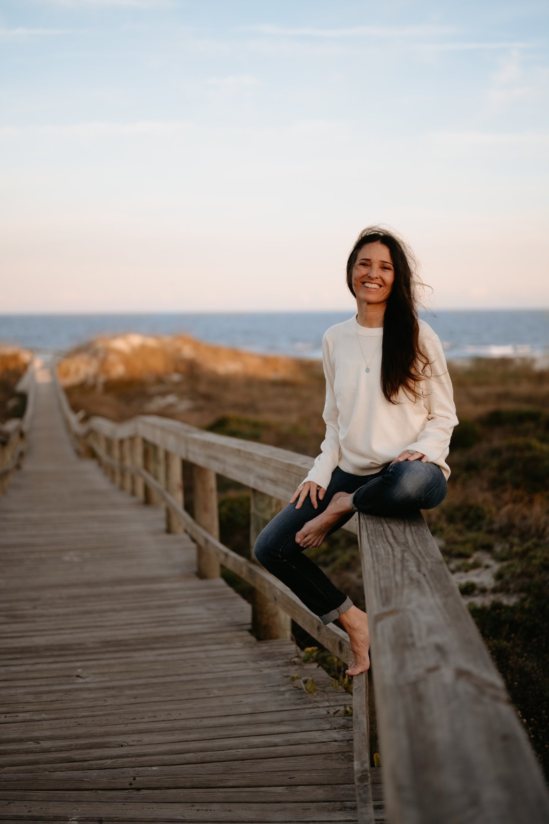 Michael Alexa Pitts smiling, sitting on the railing of a wooden boardwalk that leads over dunes to the ocean at twilight