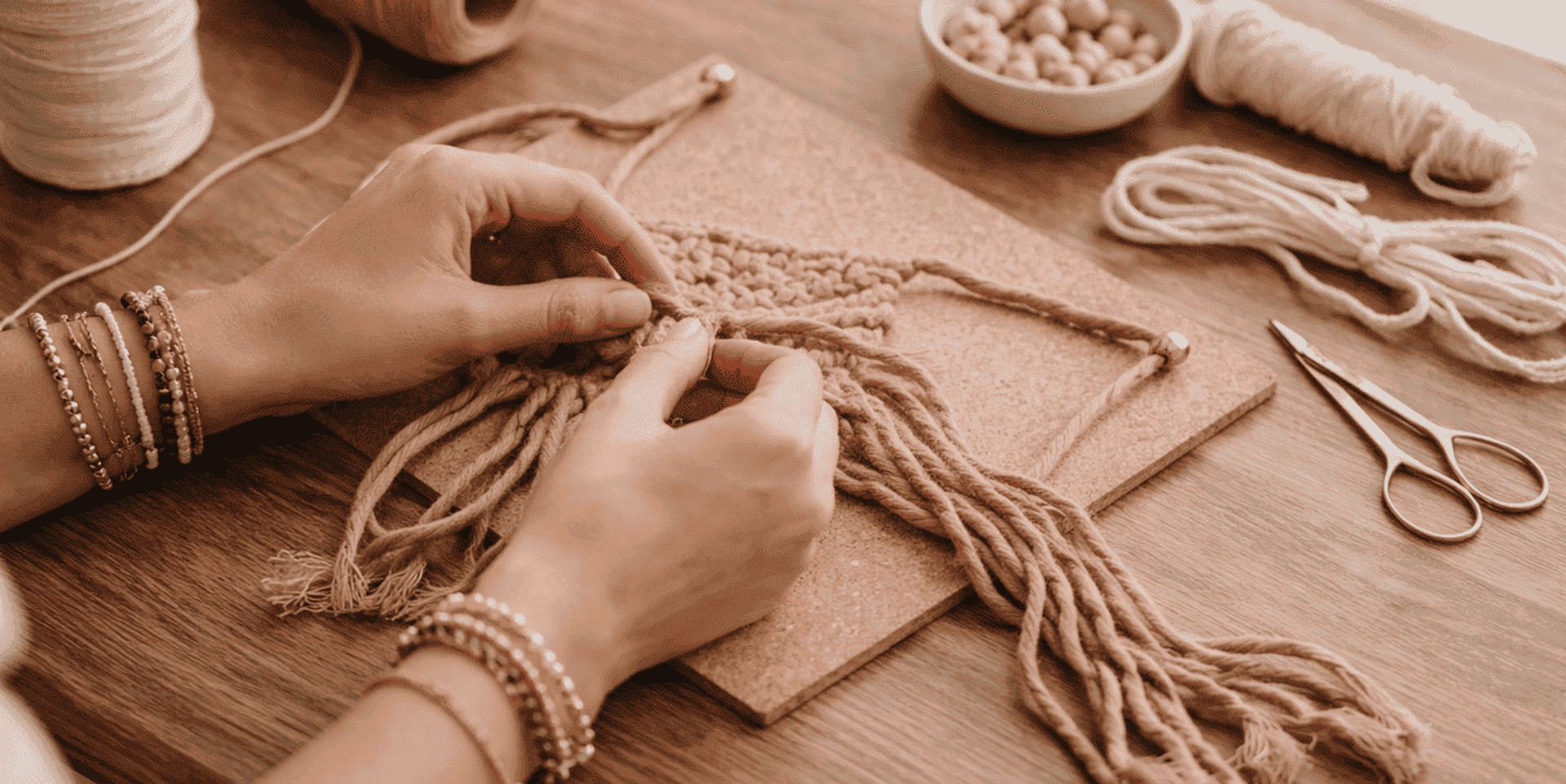 manos tejiendo macramé en colores tierra, fondo de mesa de trabajo con cuentas de madera e hilos naturales