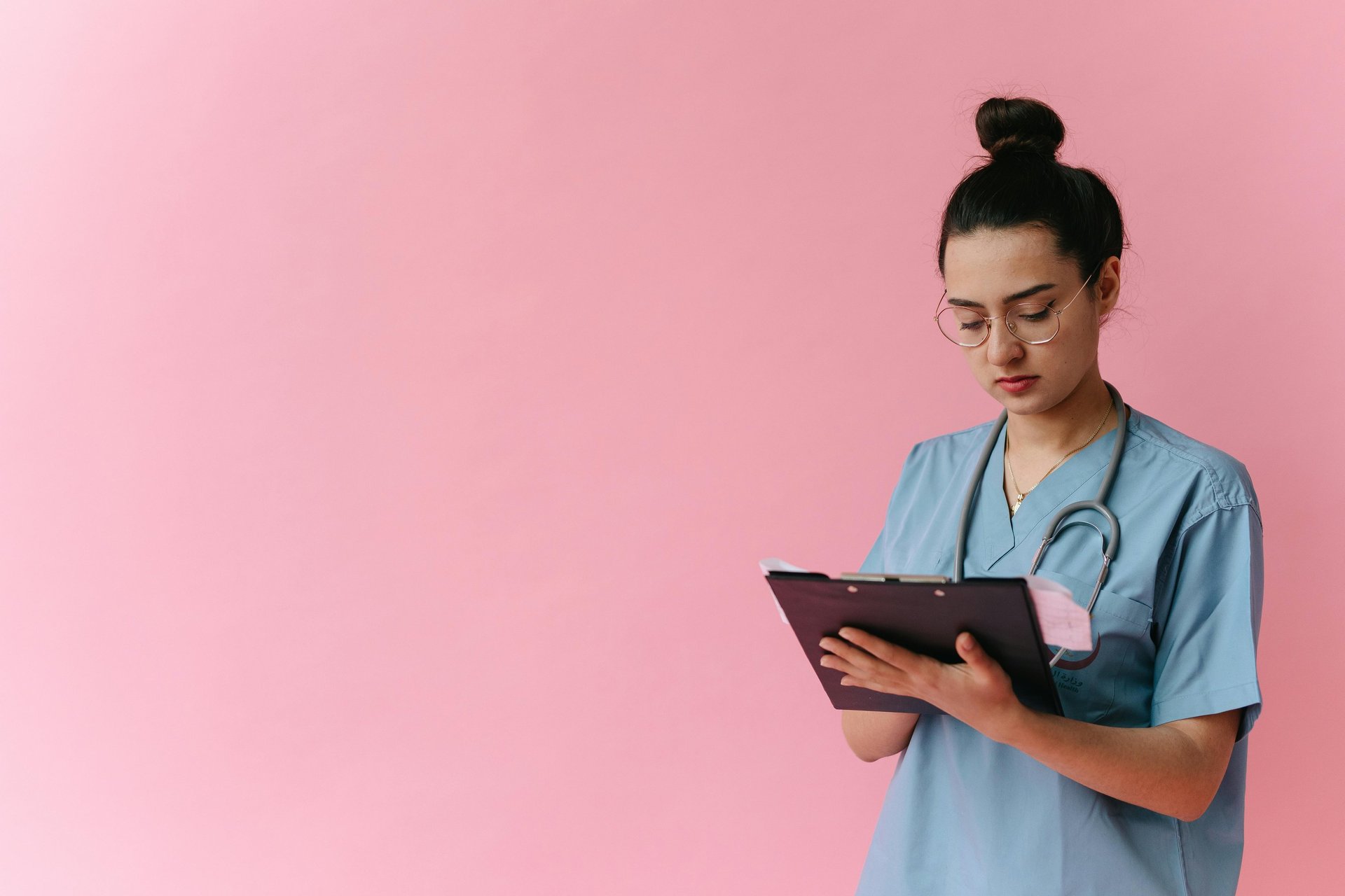 Woman in light blue scrubs, wearing glasses and looking at a folder. She has her hair in a bun