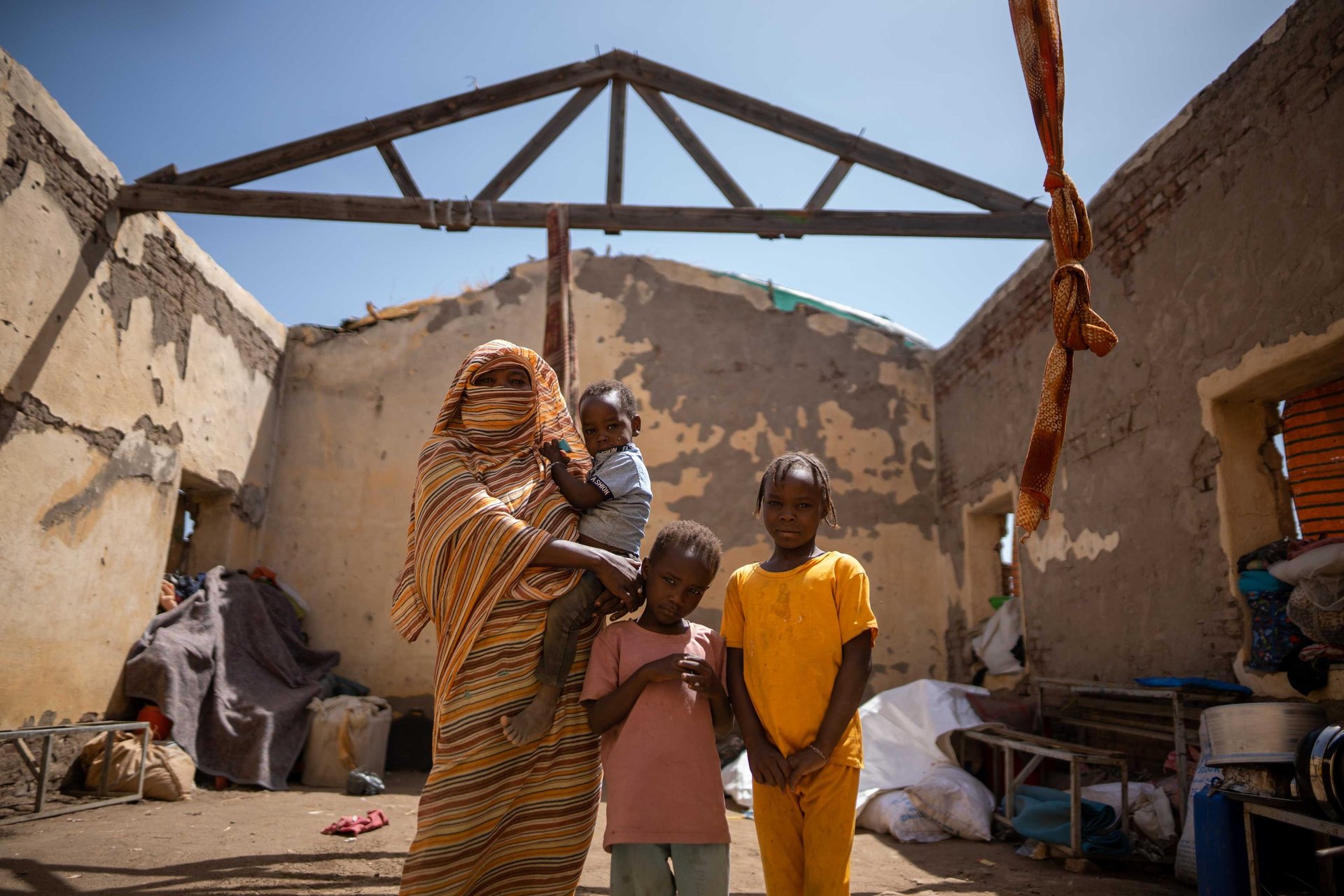 Sudanese woman and children standing inside a roofless home damaged by conflict.