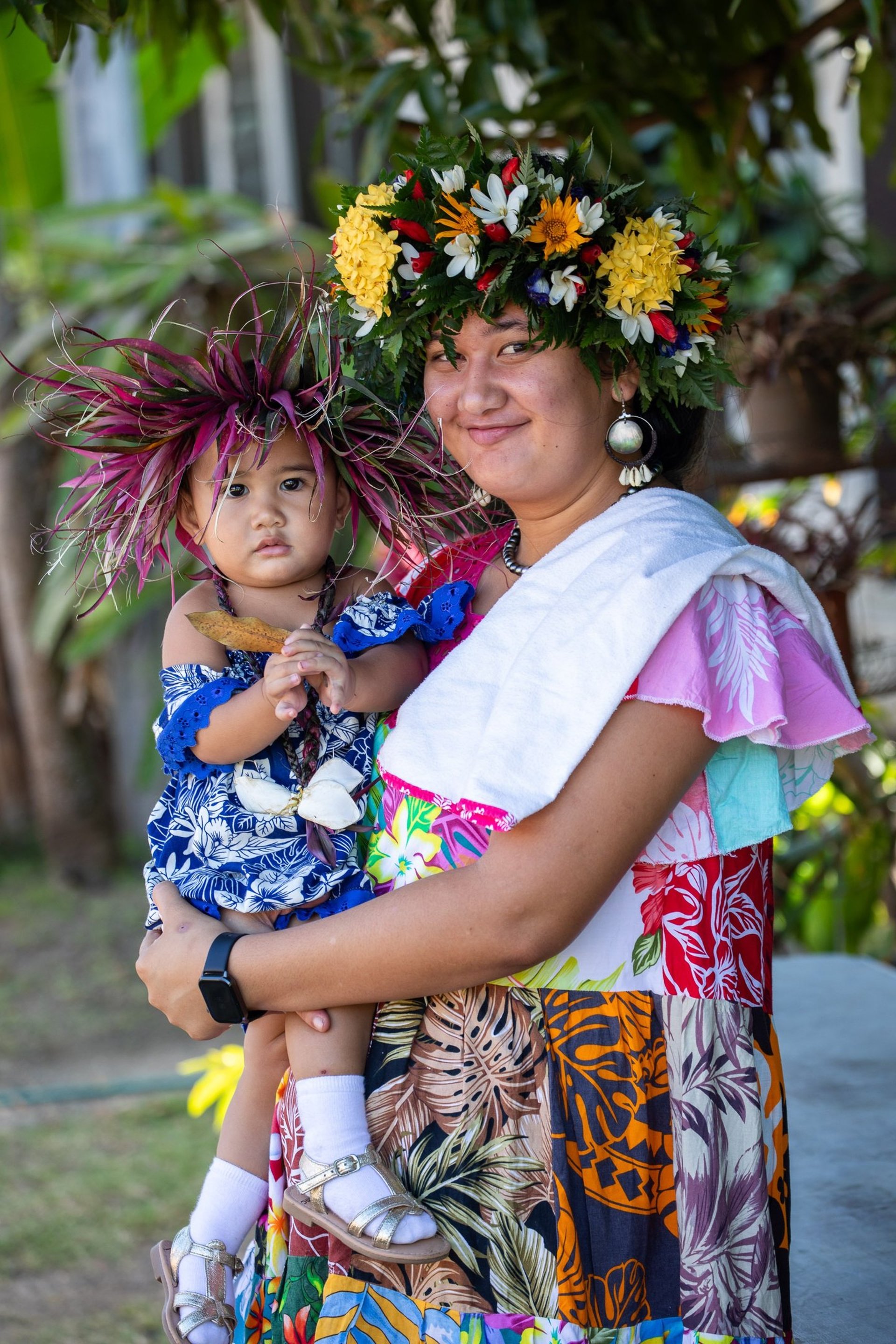 Femme polynésienne habillée en costume traditionnel qui tient son enfant dans les bras portant des couronnes de fleurs 