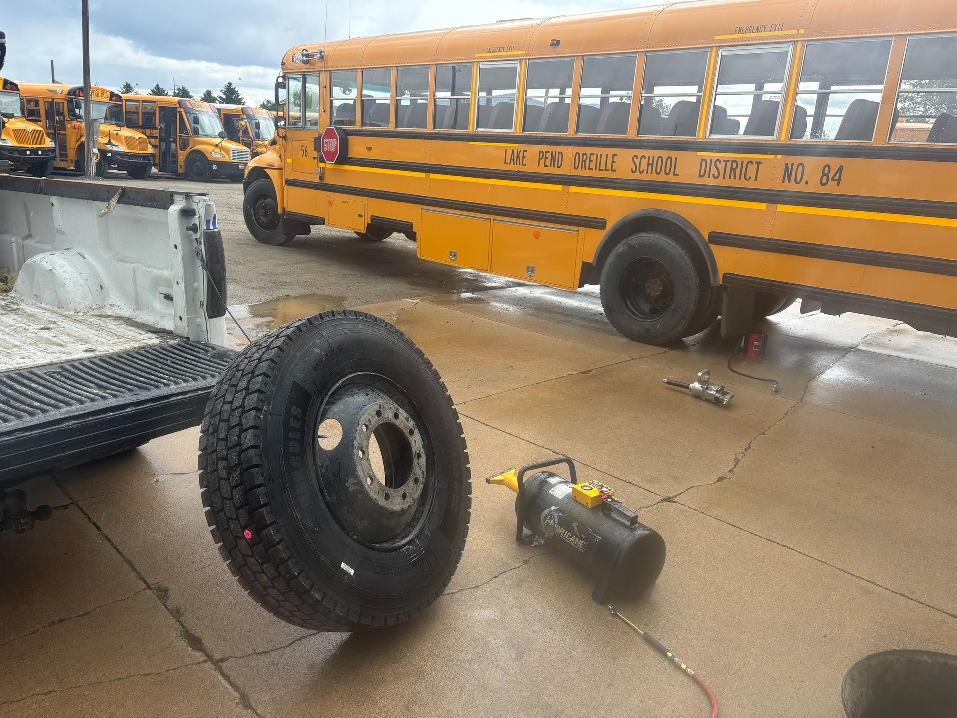 A mechanic services a yellow school bus tire and wheel on a wet pavement lot.