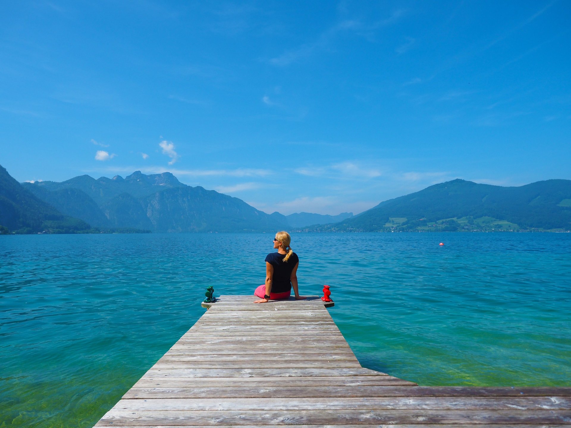 Blonde Frau sitzt am Steg an einem See und schaut in die Ferne