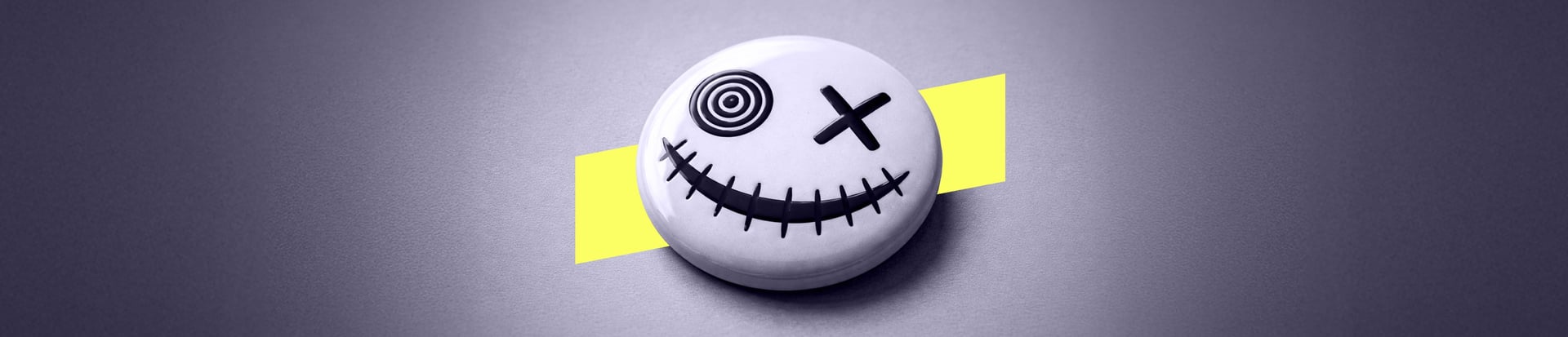 Smiling button face with one spiral eye and stitched grin on a grey background with a yellow diagonal shape.