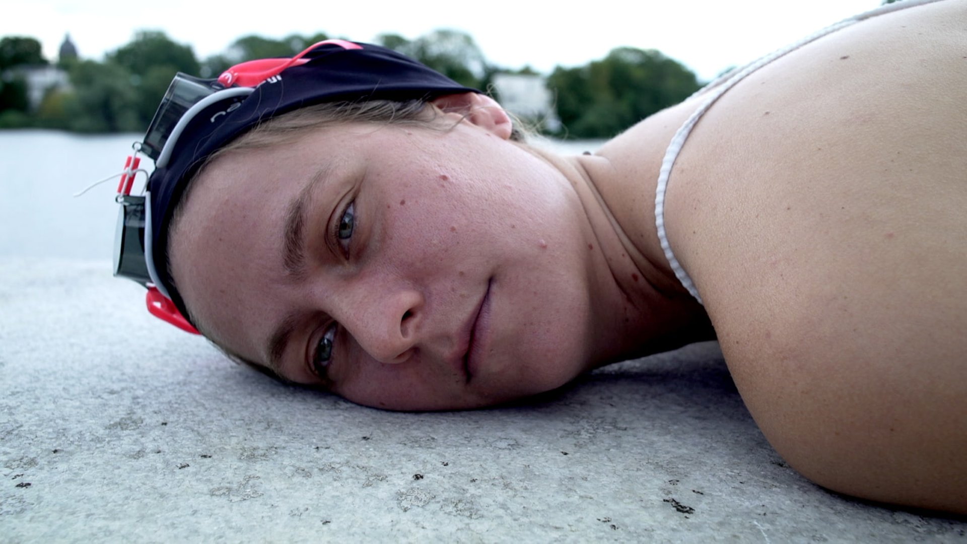 Film still from "Fear" by Tina Bikic showing a close-up of a swimmer wearing yellow goggles, crouching among tall grass.