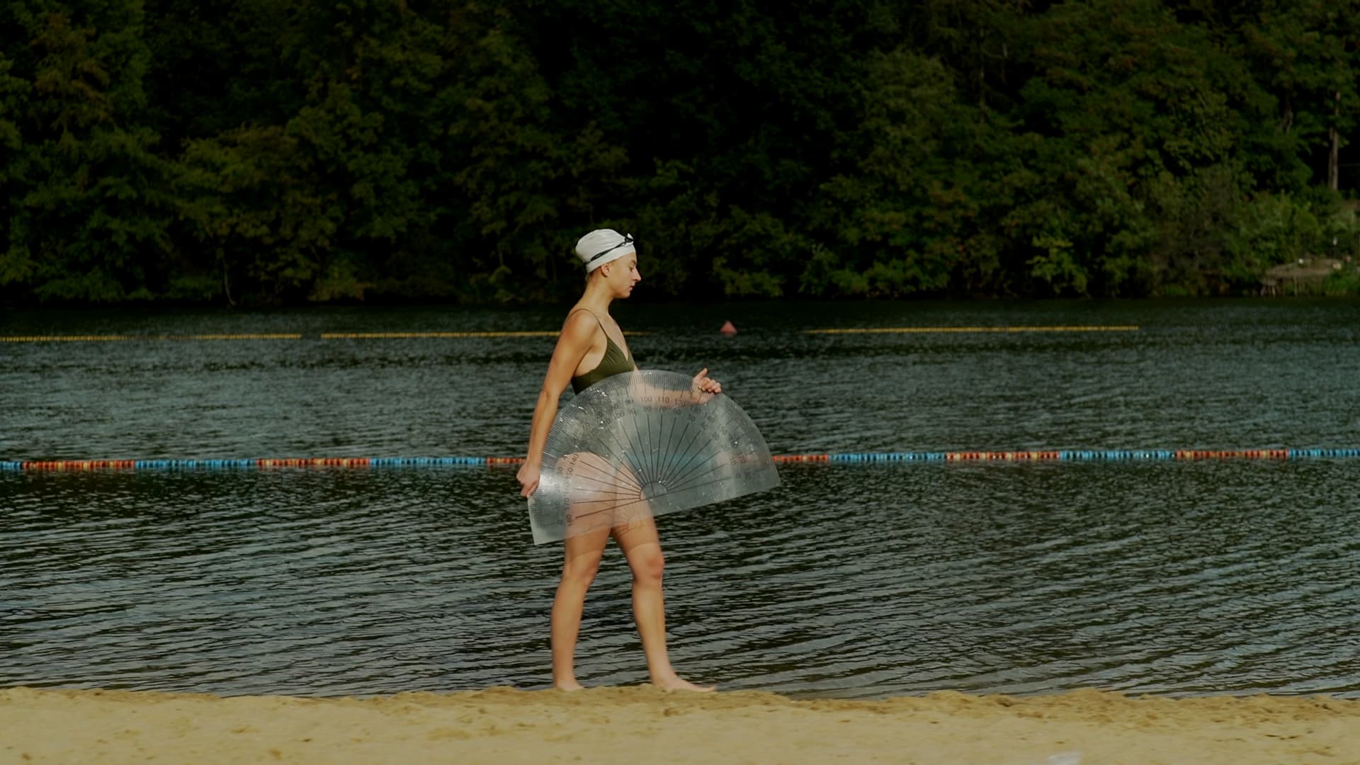Film still from "Belonging" by Tina Bikic of a woman walking along the water’s edge with forest in the background.