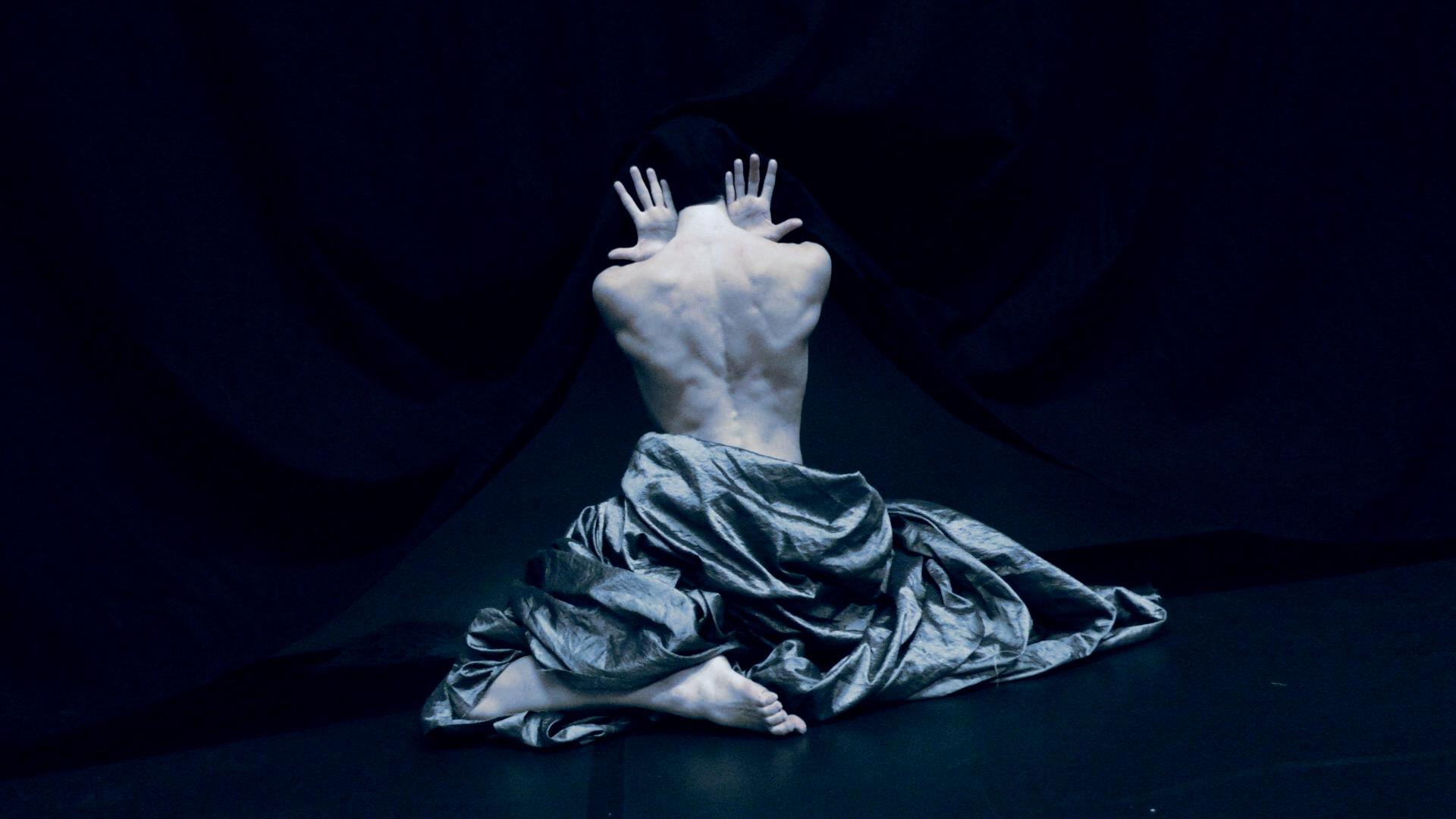 Film still from “Der Schatten” by Tina Bikic showing a dancer standing upright with a mask, body partially illuminated.