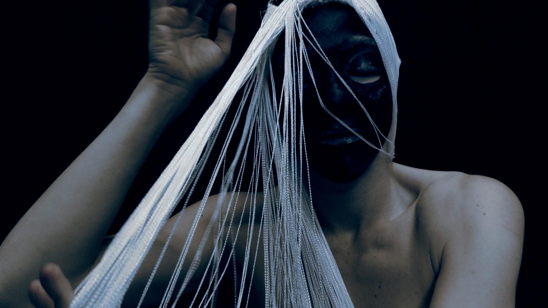 Film still from “Der Schatten” by Tina Bikic showing a dancer pulling translucent fabric over their masked face.