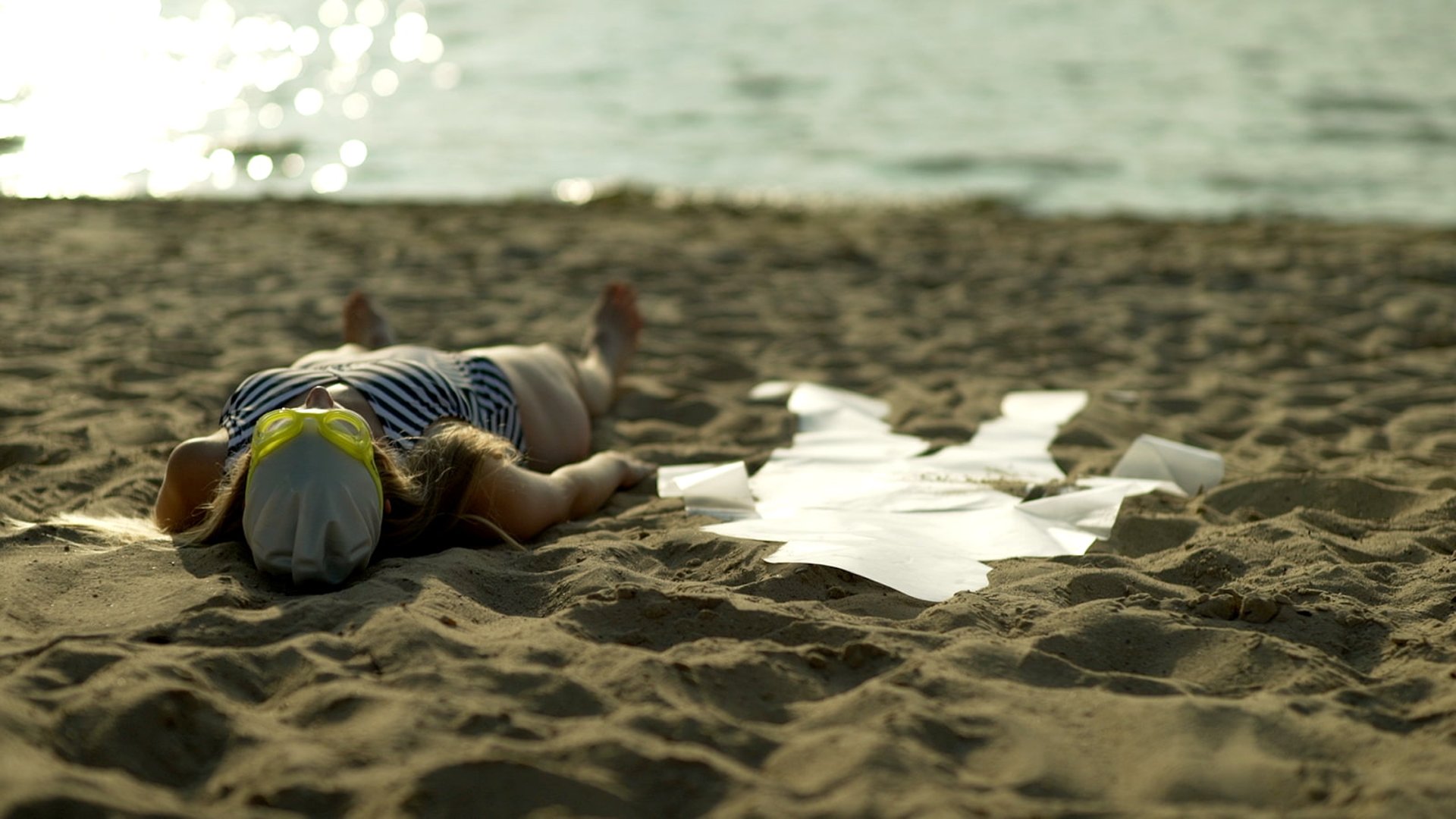 Film still from "Desire for Belonging" by Tina Bikic of a woman lying on the sand near the water’s edge with fabric.