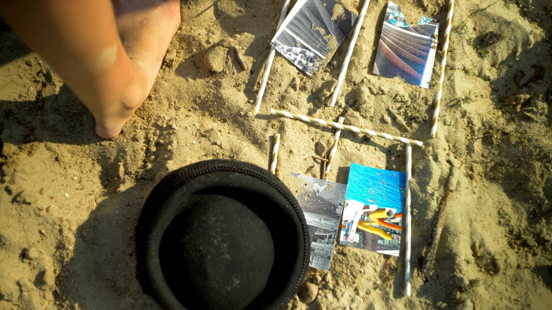 Film still from Excitement by Tina Bikic showing a black hat placed beside scattered photographs in the sand.