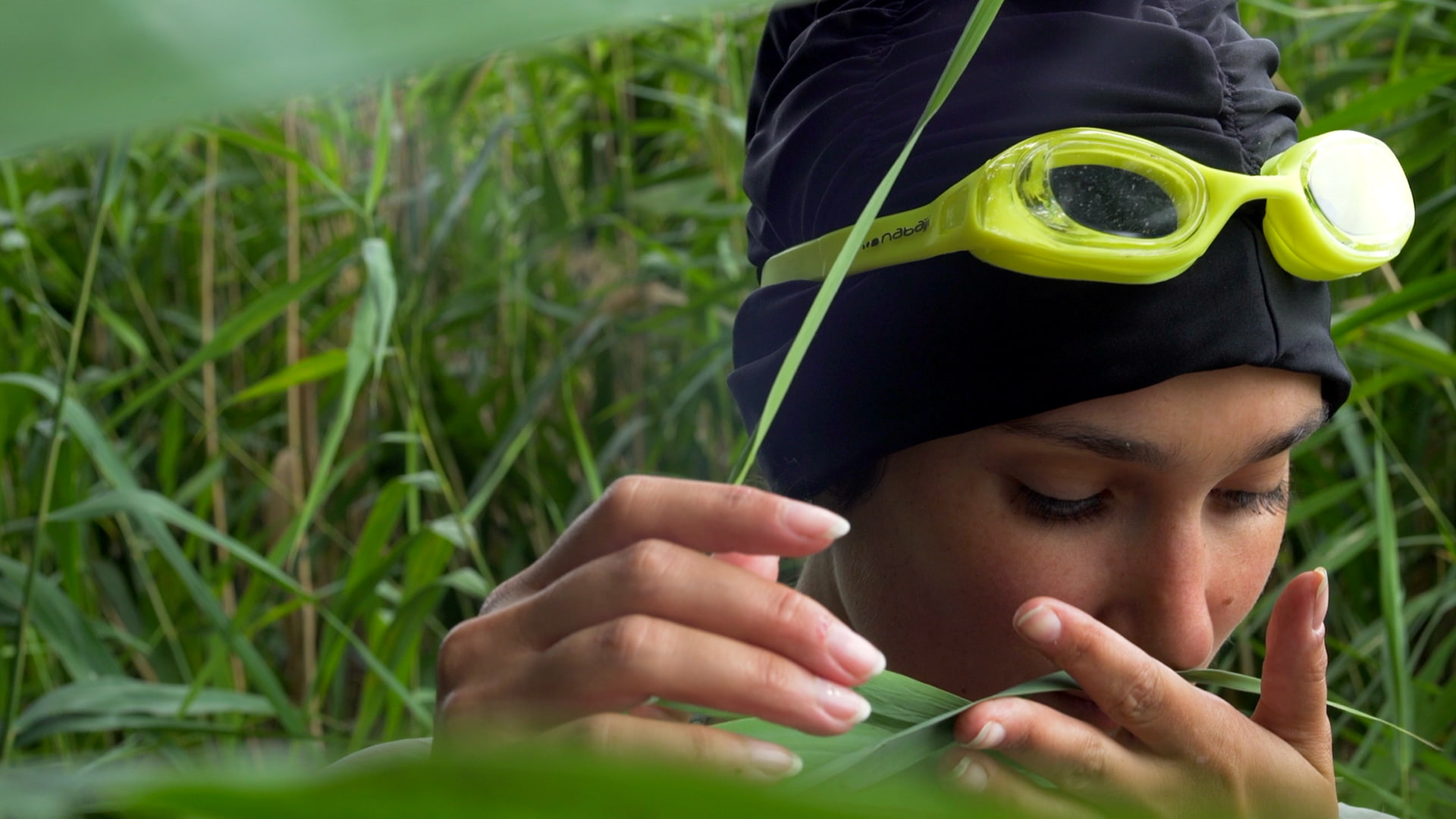 Film still from "Fear" by Tina Bikic showing a close-up of a swimmer wearing yellow goggles, smelling a tall grass.
