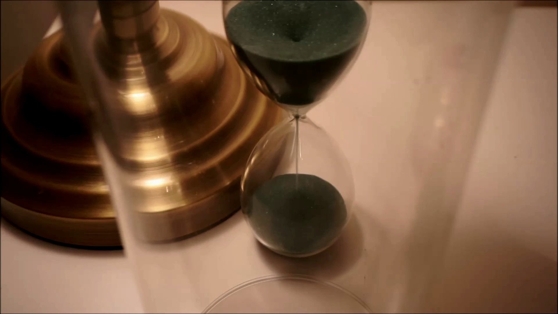 Film still from The Wait by Tina Bikic showing a close-up of a green sand hourglass beside a brass lamp base.