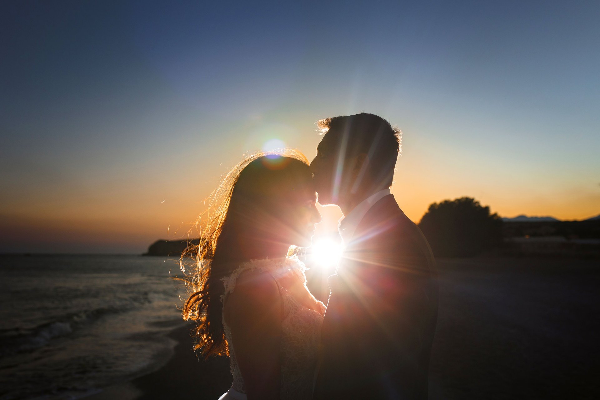 Gli sposi al tramonto si abbracciano, la luce del sole filtra tra di loro mentre lui le da un bacio sulla fronte
