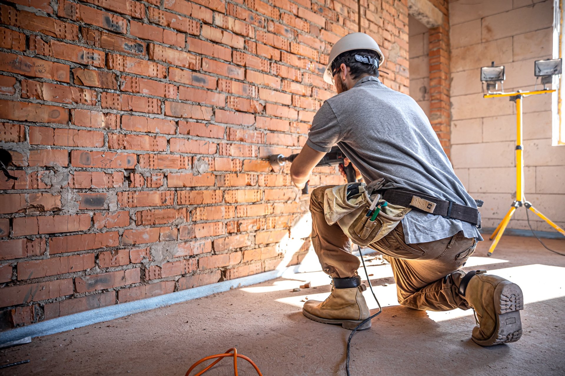 Construction worker, wearing a hard hat and safety boots, drilling a brick wall with a perforator