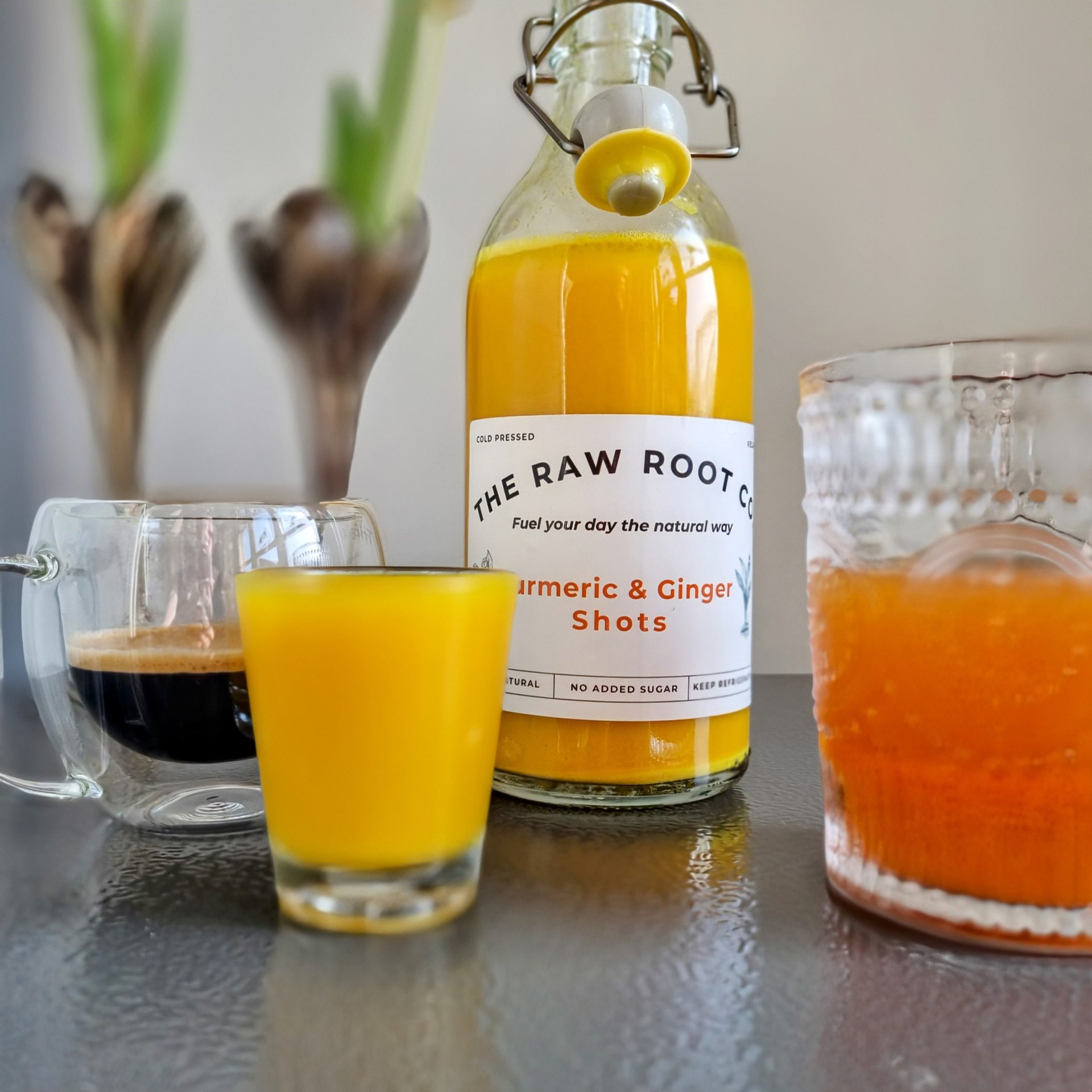 Turmeric & Ginger shot bottle with a shot glass full of Turmeric and Ginger next to an espresso coffee. The Raw Root Co.
