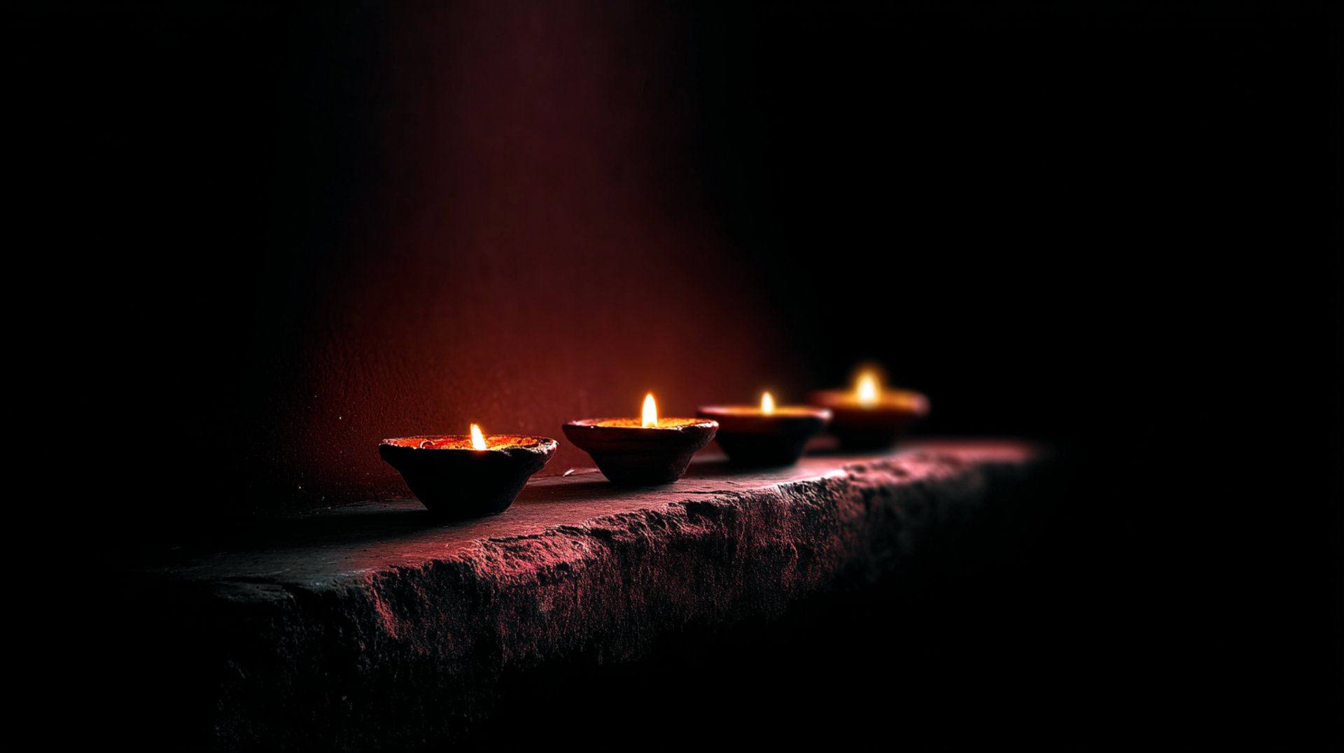 Four traditional oil candles, their small flames casting a warm, atmospheric glow against a dark, red-black background.