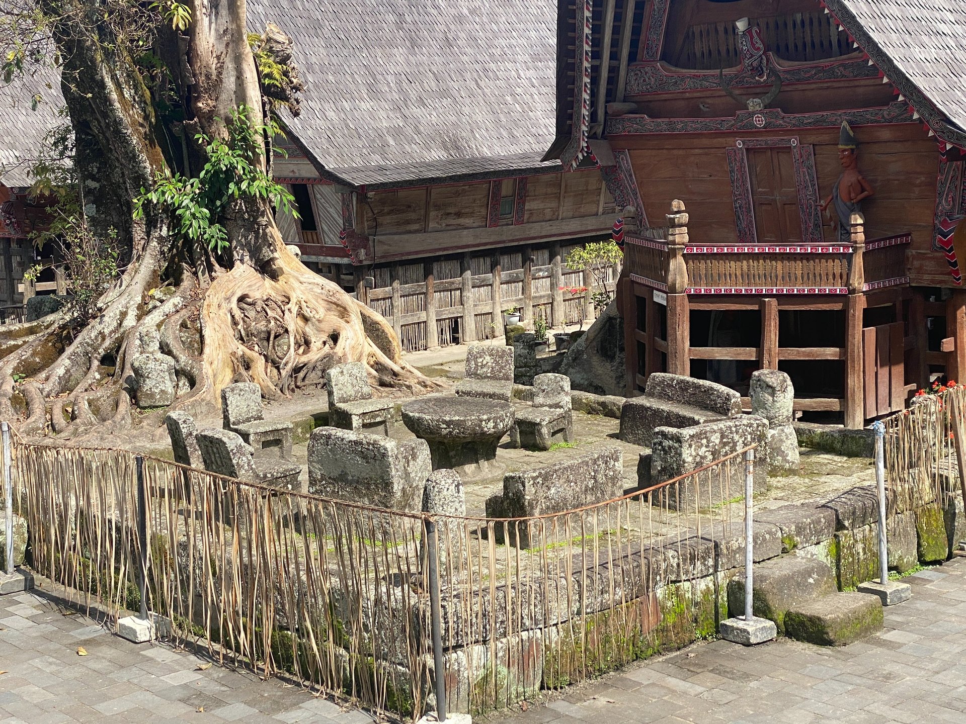 an ancient Batak court area with carved solid stone as seating with an old tree behind it and replica housing