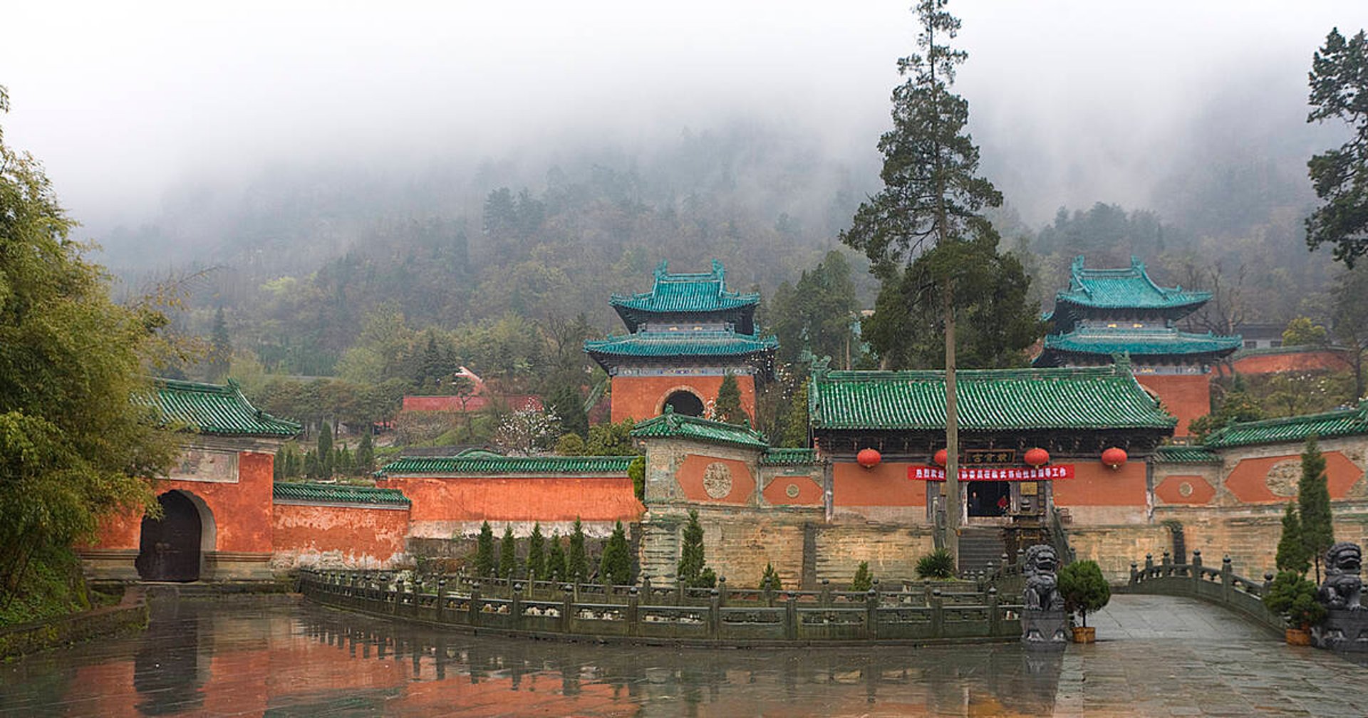 Ancient Wudang temple complex with green roofs, misty mountains, and a stone bridge over water.