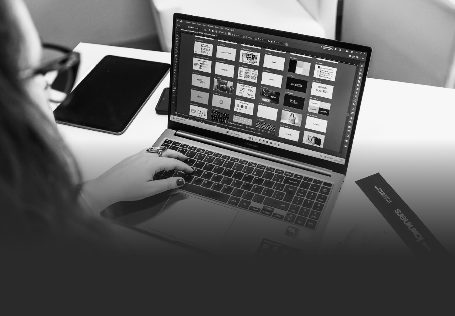 Moça digitando em notebook aberto com projeto de id visual em mesa de trabalho branca.