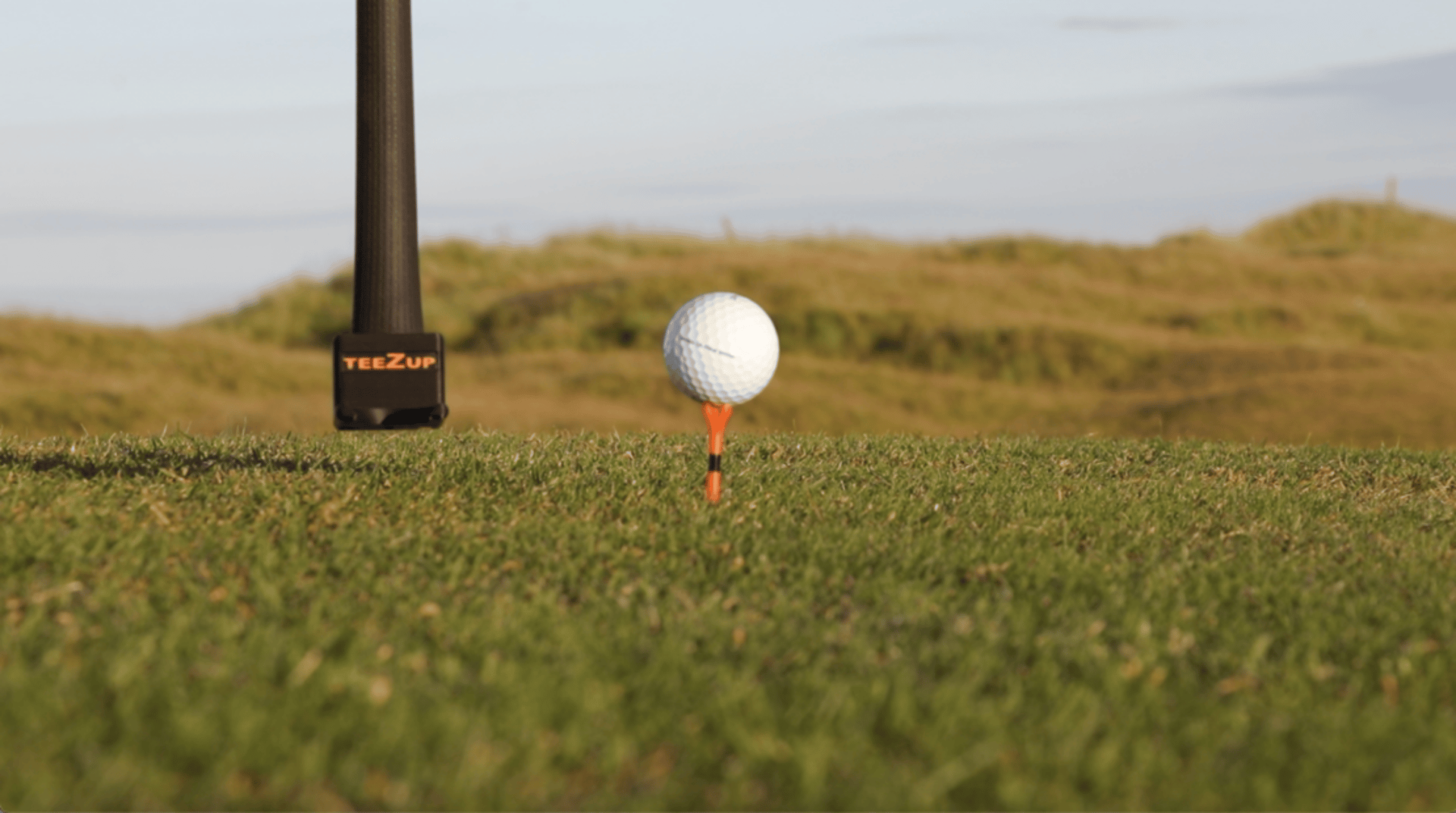 A golf ball on an orange tee with a 'TeeZUP' tool positioned beside it on a grassy course, with hills in the background.as