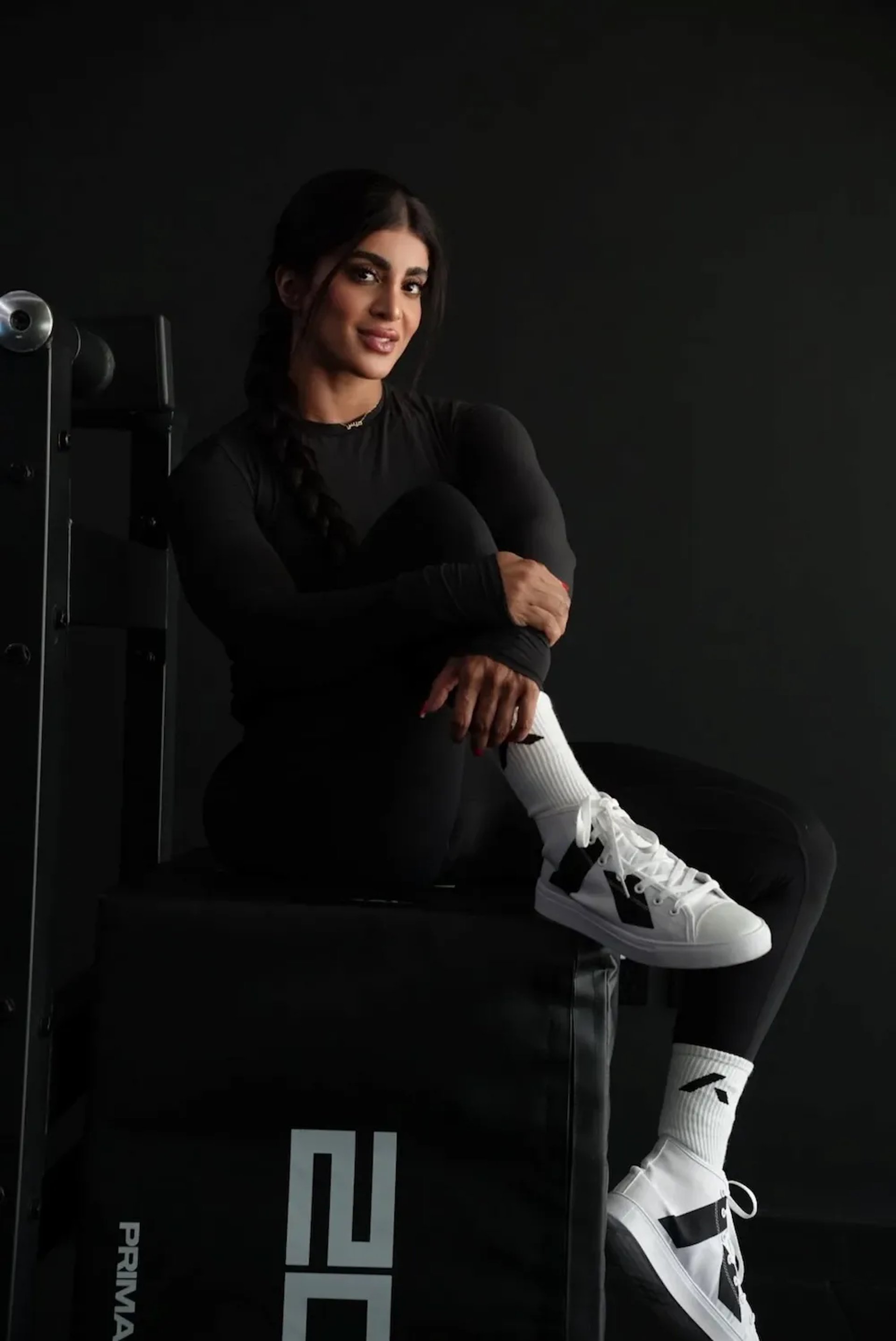 Mais Alhajri in black sportswear and white sneakers sitting in a gym setting, fitness and strength training concept."