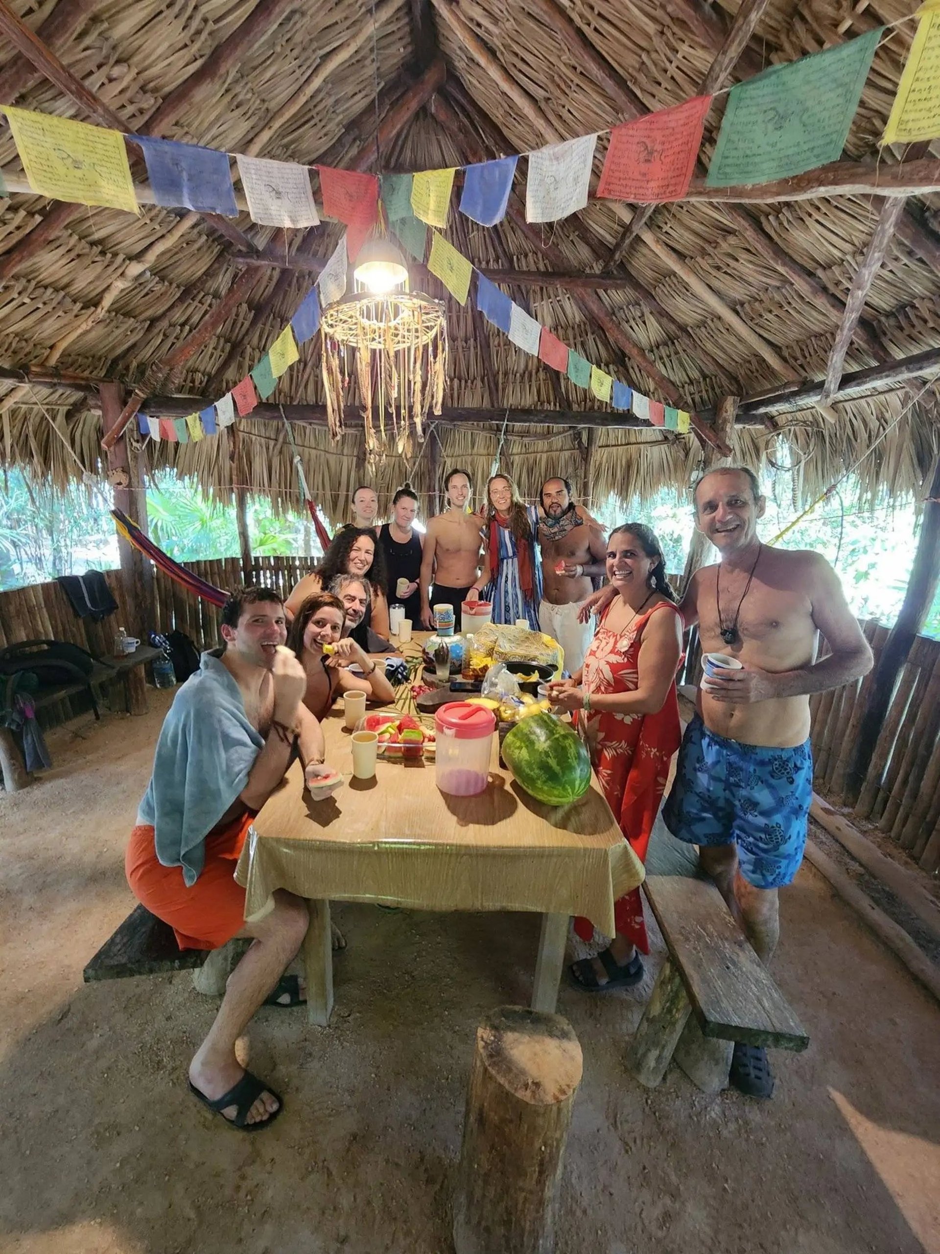 Un grupo de amigos disfruta de una comida tropical juntos dentro de una cabaña con techo de paja decorada con banderas.