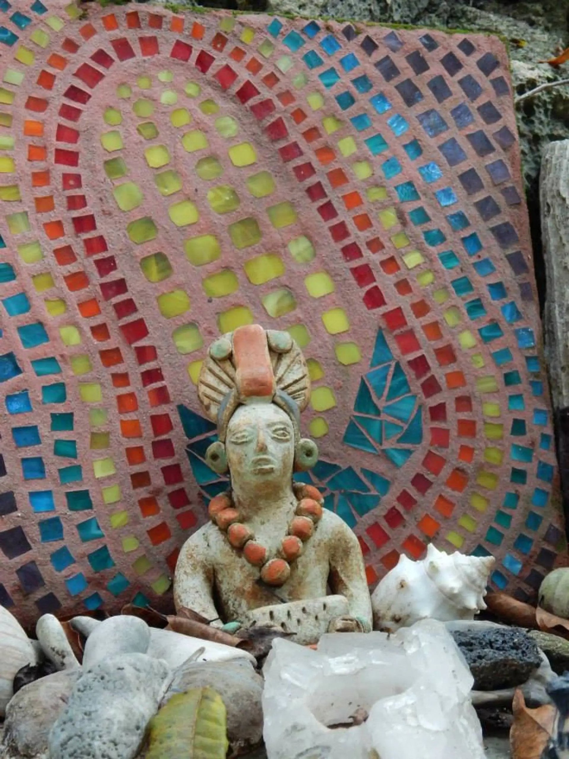 Una estatua maya de arcilla se encuentra frente a una colorida pared de mosaico con piedras y conchas marinas.