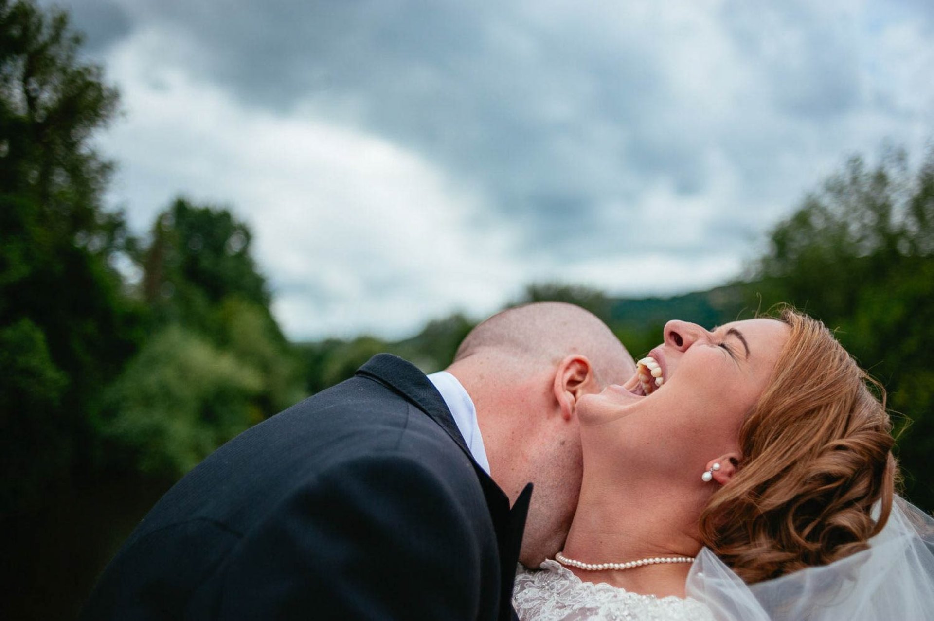 Bride laughing as groom embraces her at outdoor South Wales wedding, captured in candid documentary style by Owen Mathias