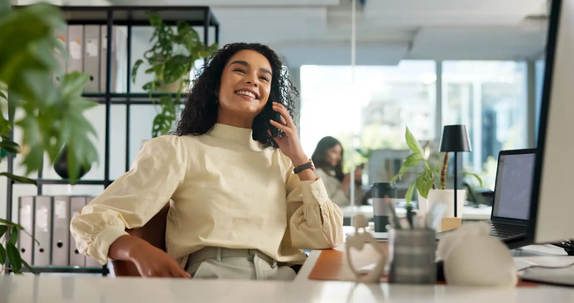 Smiling woman talking on the phone at a modern office desk with a laptop, plants, and office supplies