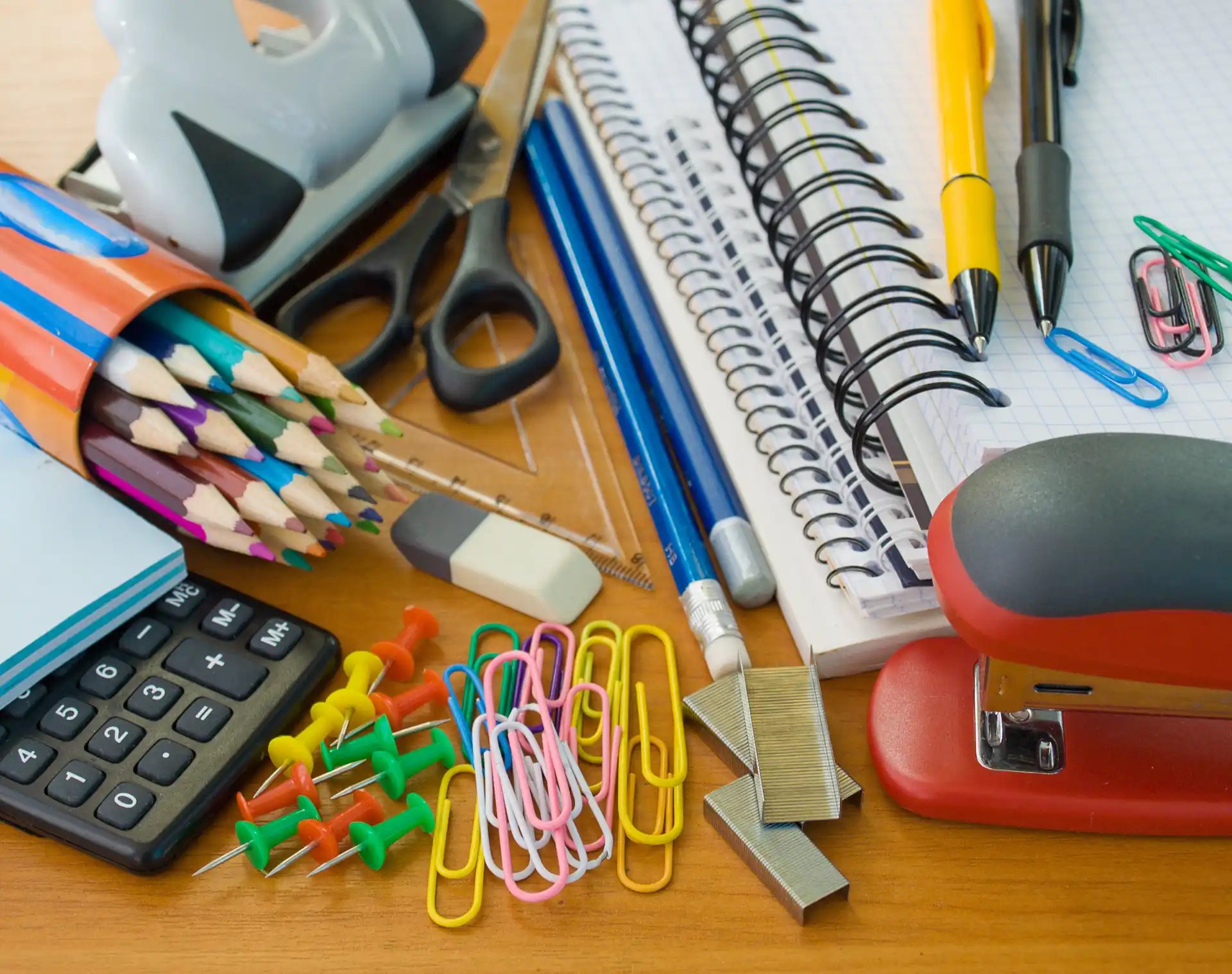Office supplies on a wooden desk, including stapler, notebooks, pens, scissors, paper clips, push pins, and calculator