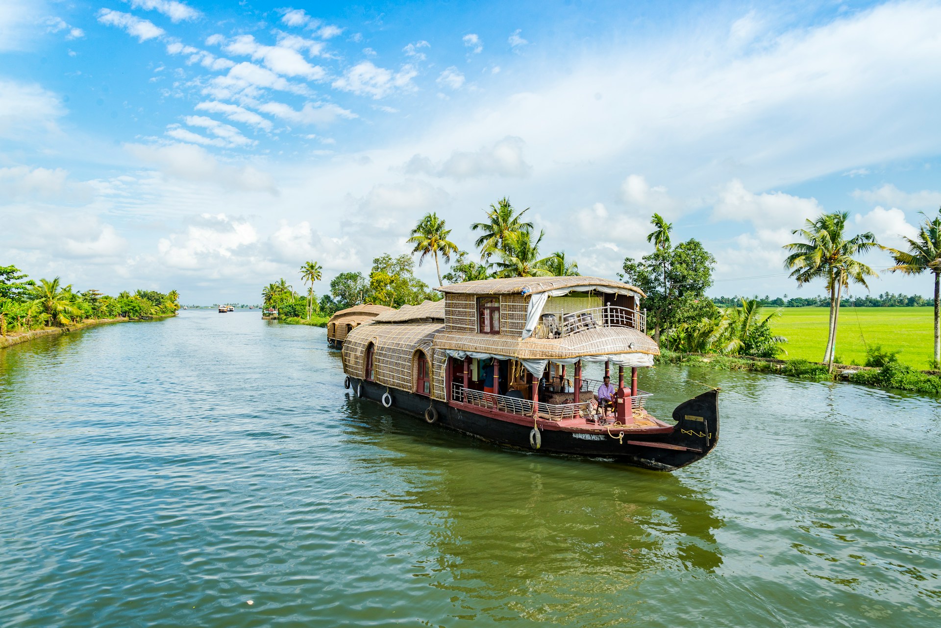 Houseboat cruising through Kerala's backwaters, surrounded by lush palm trees and vibrant greenery under a blue sky.