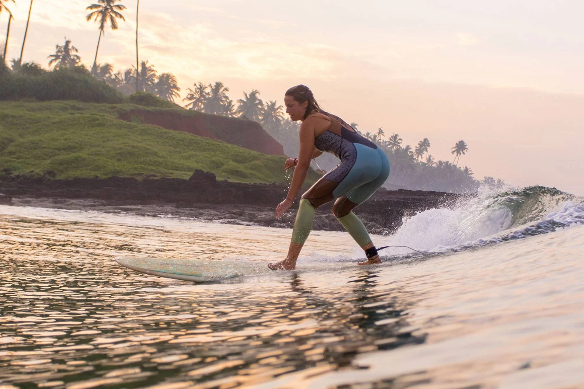Girl surfing on a wave in Varkala, Kerala, with palm trees and cliffs in the background during sunrise.