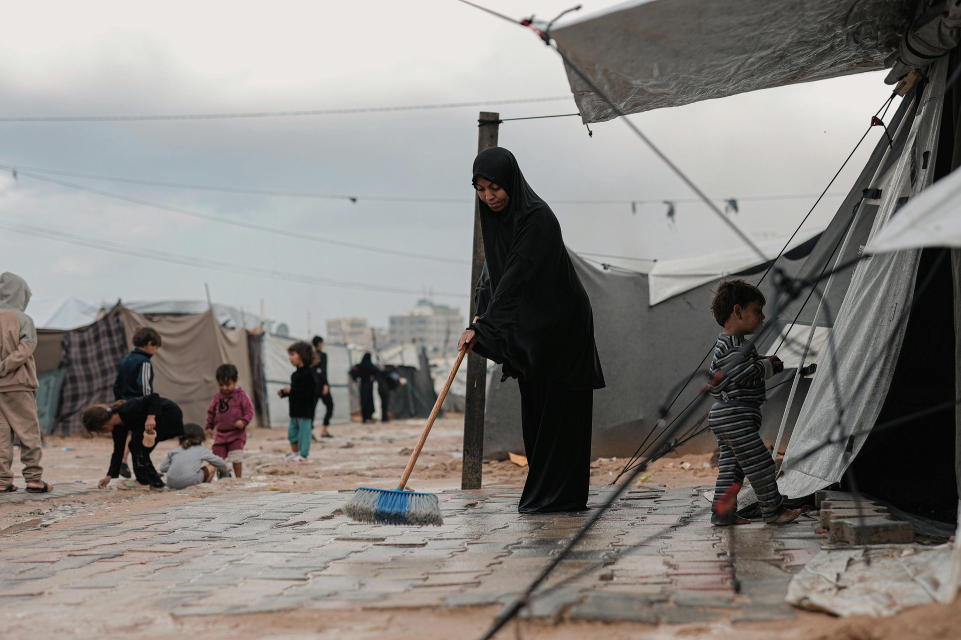 femme réfugiés gaza devant sa tente en train de nettoyer le sol, ses enfants jouent à l'extérieur dans la boue
