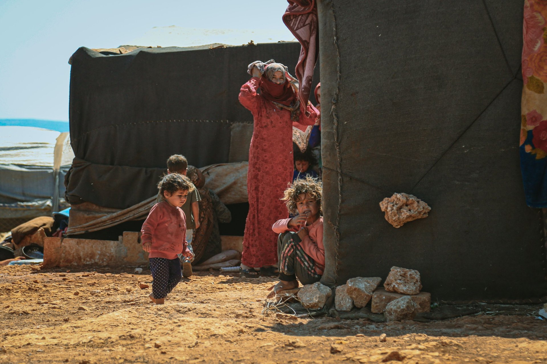 femme réfugiés gaza devant sa tente en train de nettoyer le sol, ses enfants jouent à l'extérieur dans la boue
