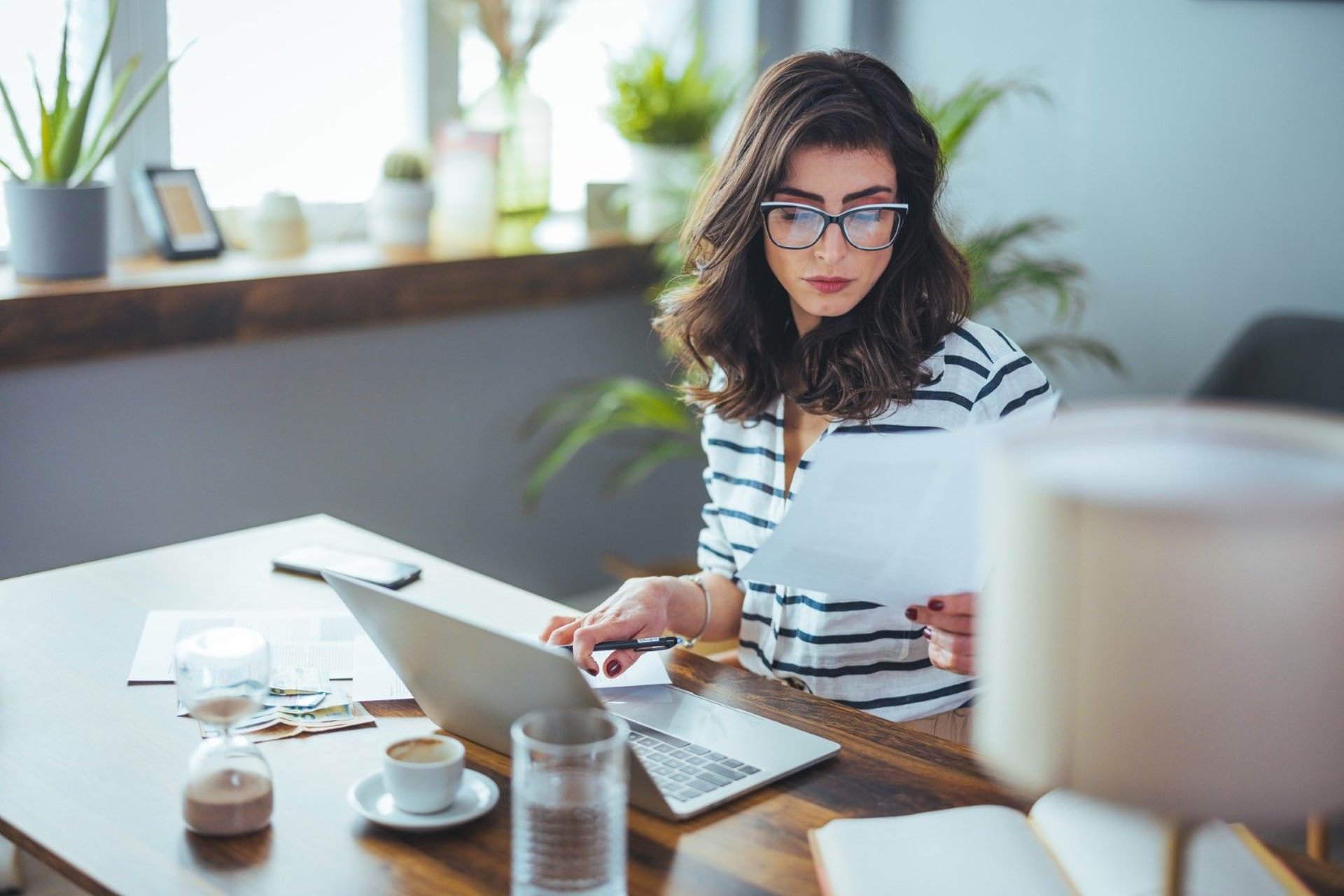 A businesswoman in glasses is seated at a desk with a laptop, appearing anxious about her financial challenges and debts.