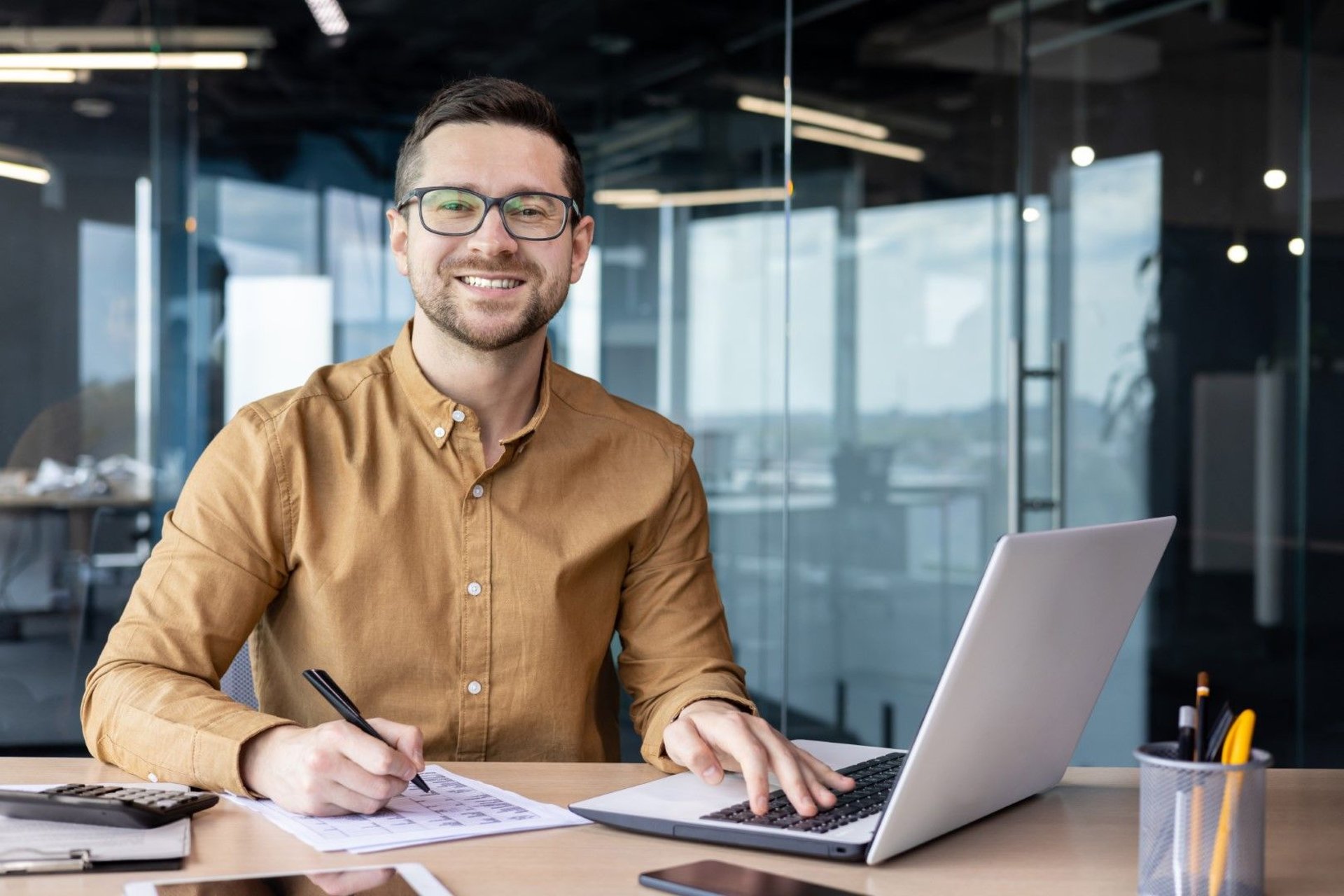 Portrait of a cheerful man in a shirt at his desk, working on a laptop with financial documents and a calculator nearby.