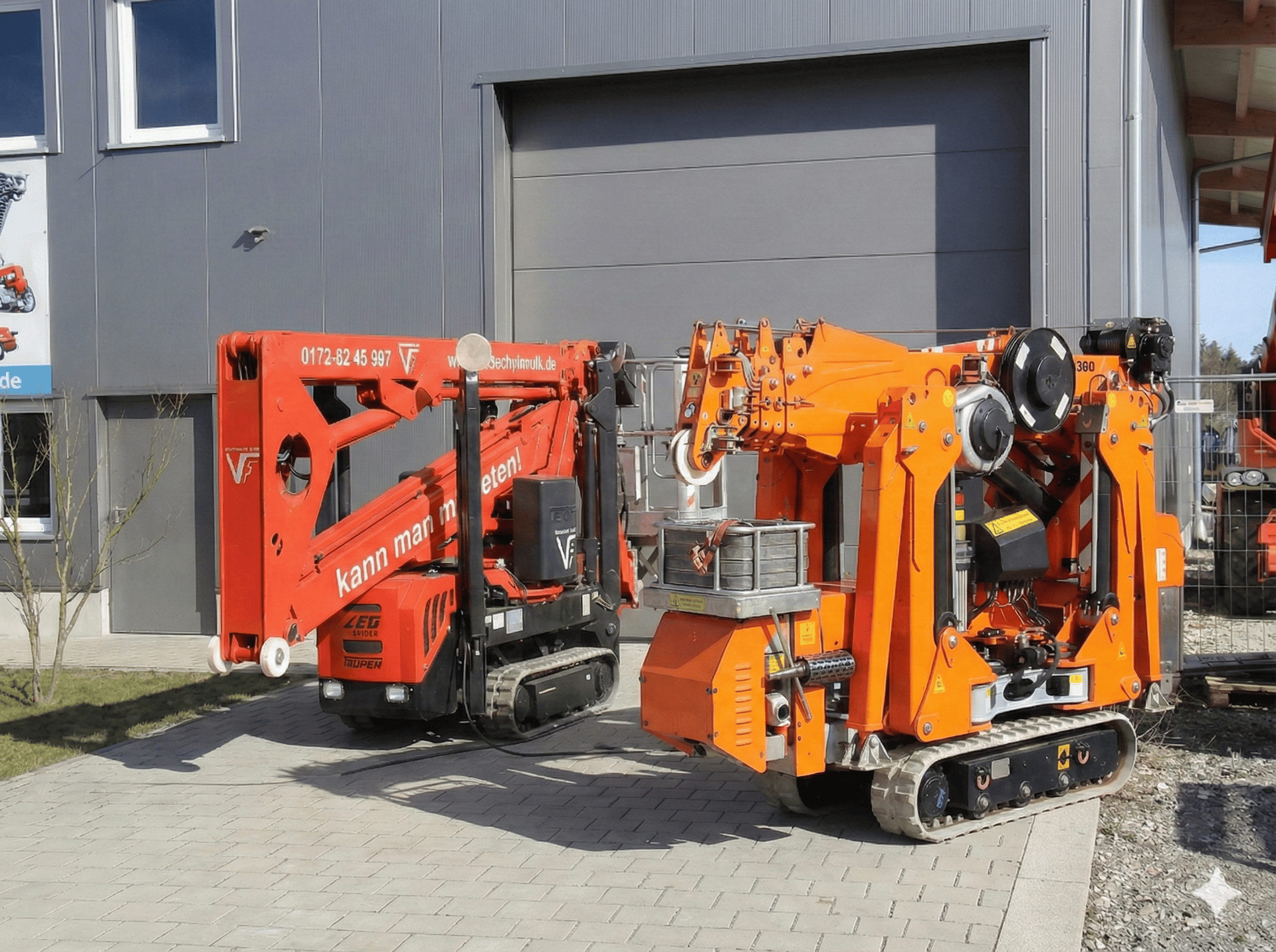 Eine rote Raupen-Arbeitsbühne und ein oranger Minikran stehen zusammengefaltet nebeneinander auf einem gepflasterten Hof.