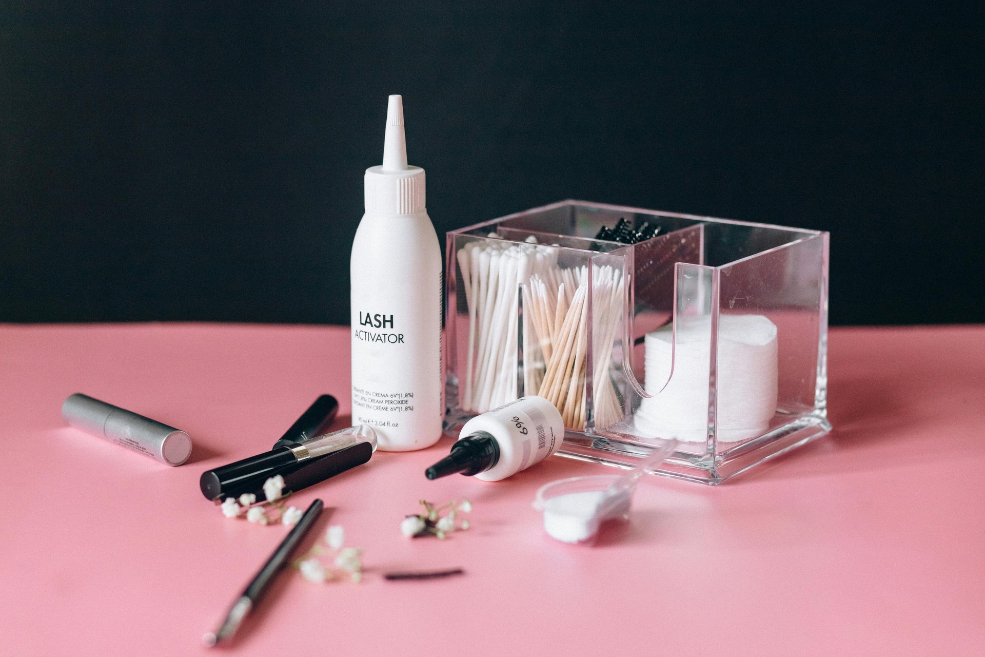 Productos profesionales para lashes sobre base rosa con fondo rosa y negro, junto a insumos desechables para aplicación.