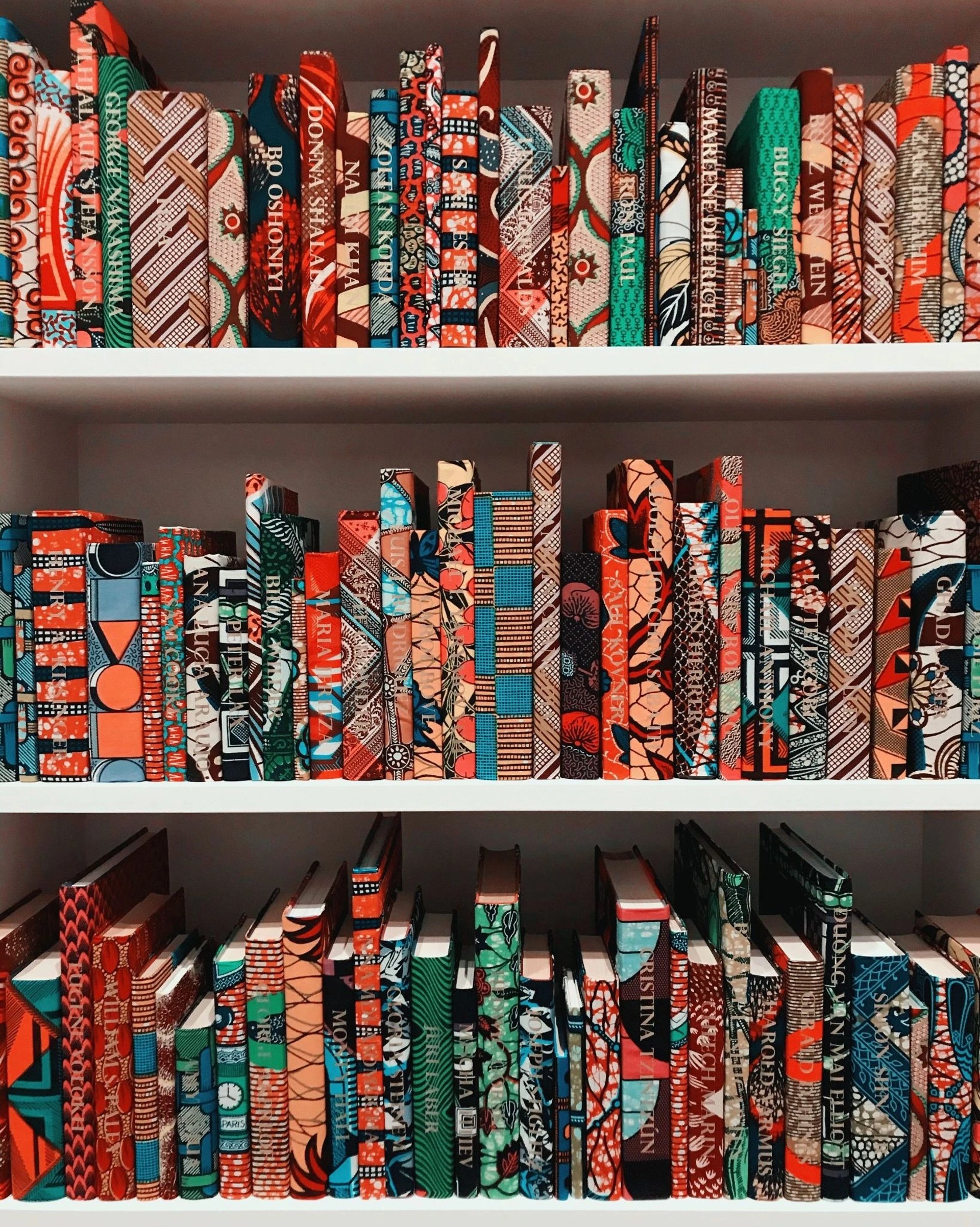 Une étagère chargée de livres présentant des motifs colorés et diversifiés de style wax sur leurs couvertures.